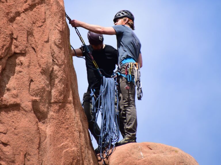 Two climbers with helmets and gear ascending a red sandstone cliff under a clear blue sky.
